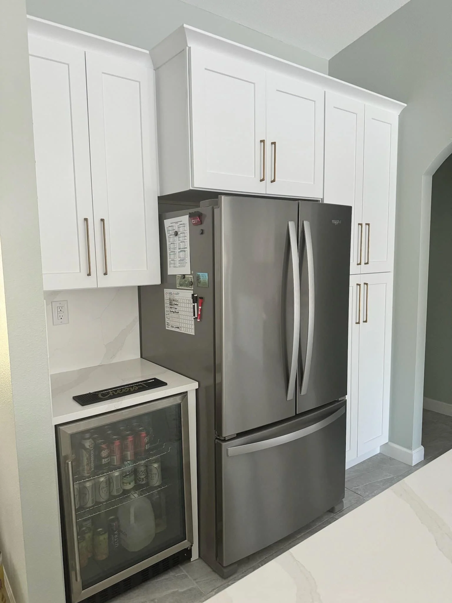 Stainless steel refrigerator next to white kitchen cabinets and a small beverage fridge with cans inside.