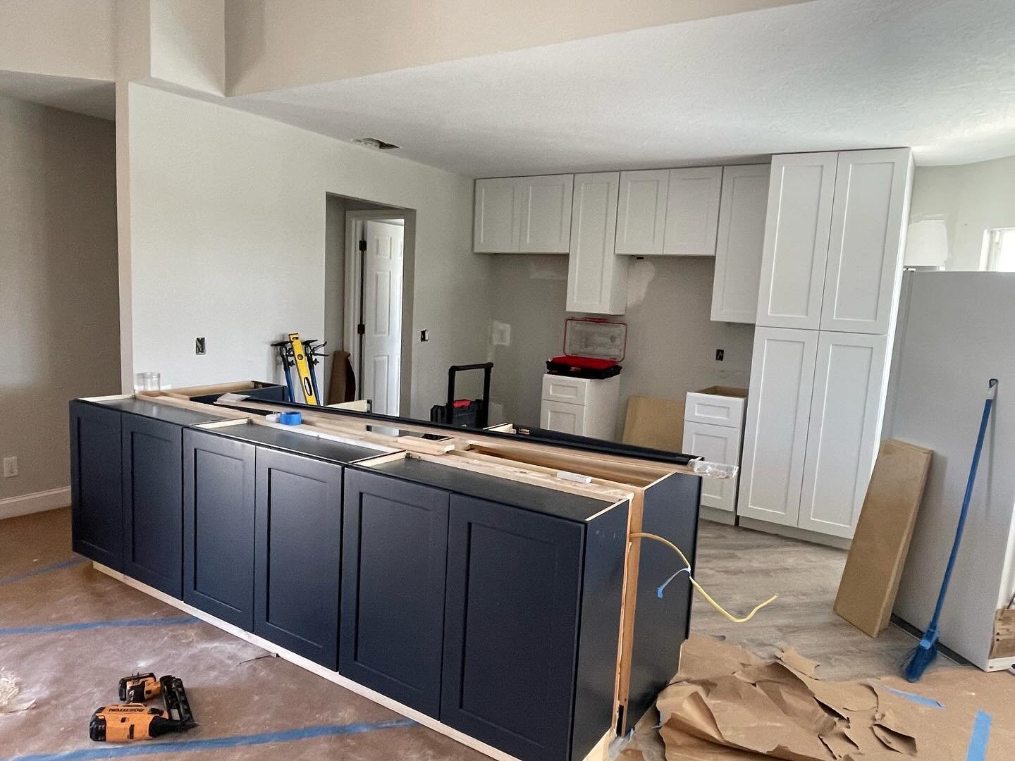 Kitchen under renovation with dark blue cabinets installed in the island, white cabinets on the walls, construction tools and materials scattered around, and unfinished flooring.