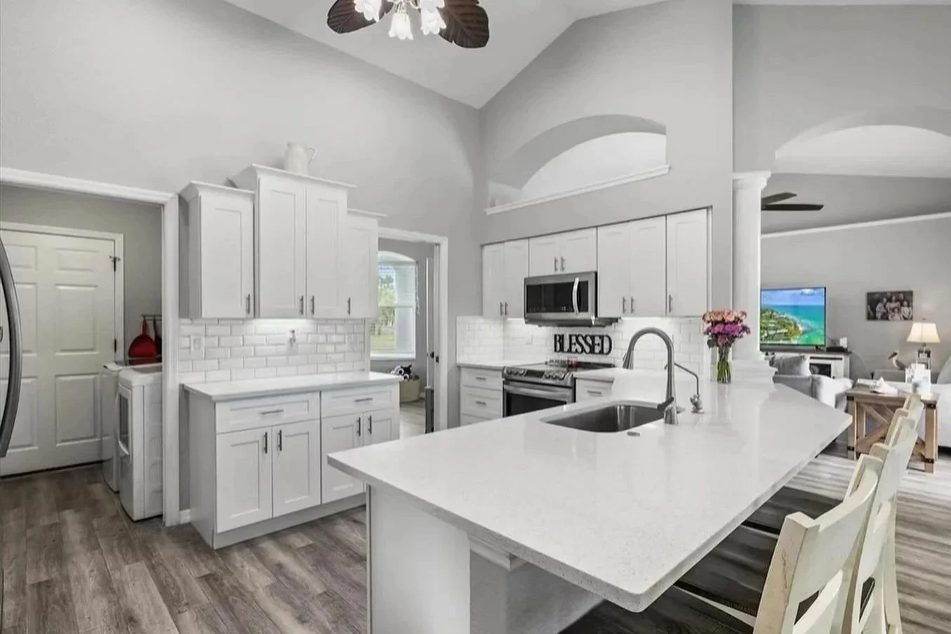 Modern white kitchen with island, stainless steel appliances, and living area with TV in the background.