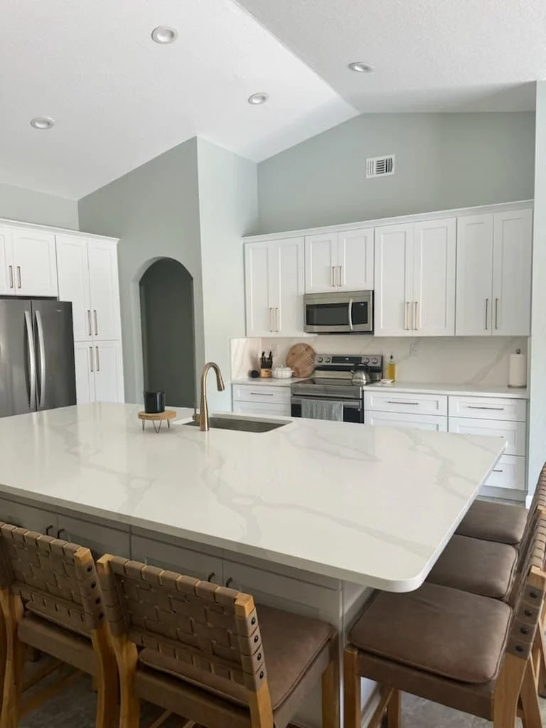 Modern kitchen with white cabinetry, stainless steel appliances, a large white island with seating, and a light gray wall.