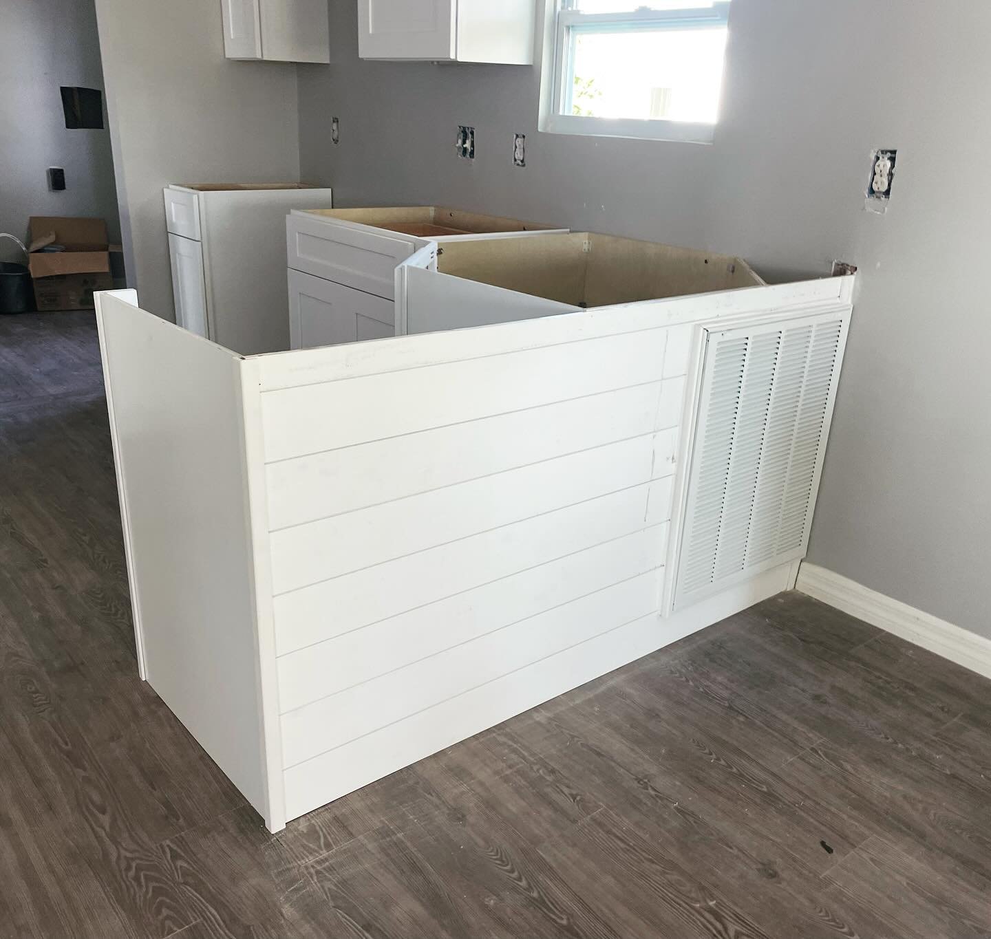 A kitchen under renovation with a white painted shiplap-style kitchen island and partial cabinetry, wooden flooring, and a window providing natural light.