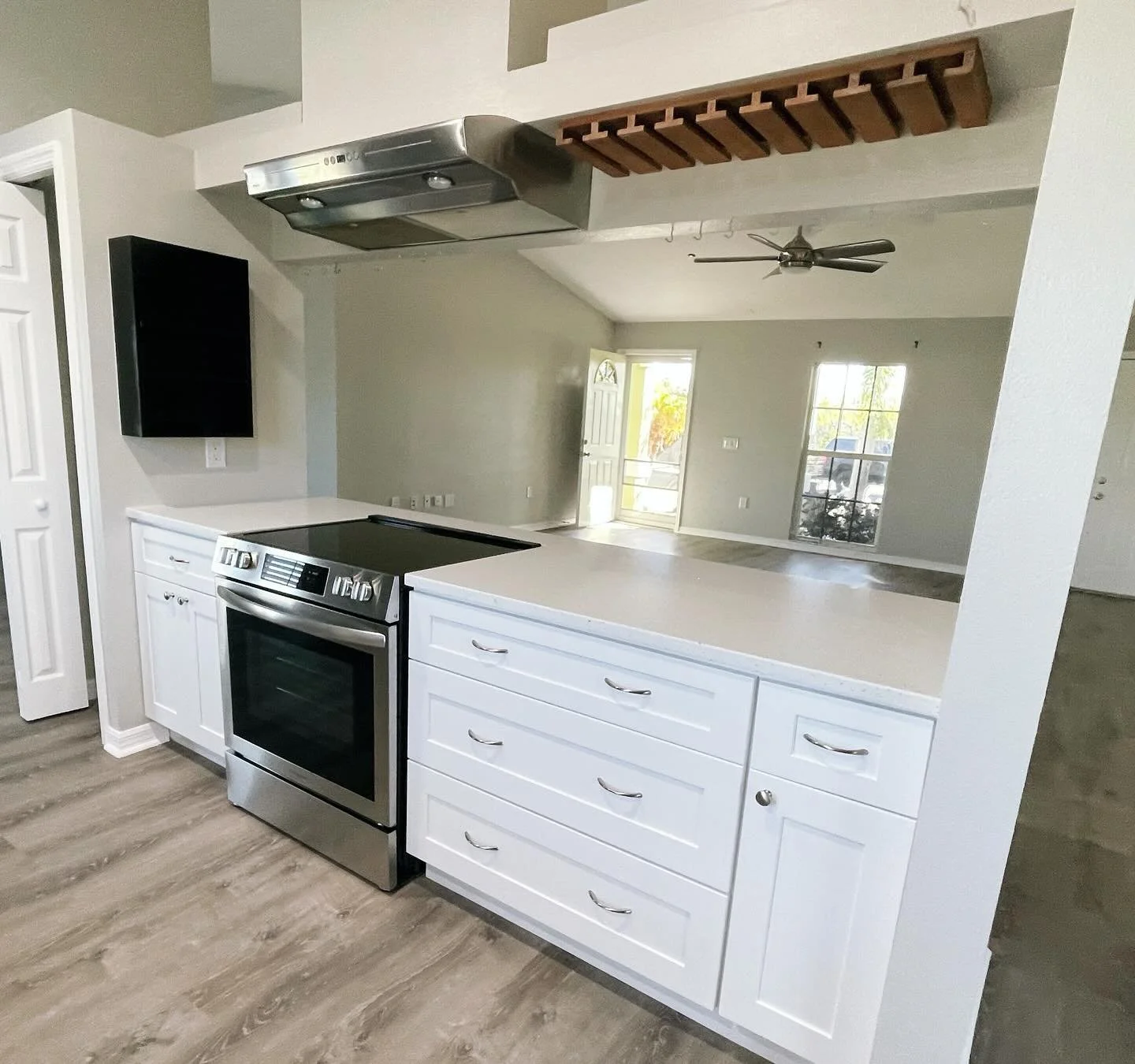 Modern kitchen with white cabinets, black and stainless steel stove, white countertops, and hardwood floors. Open view to living area with large windows and a ceiling fan.