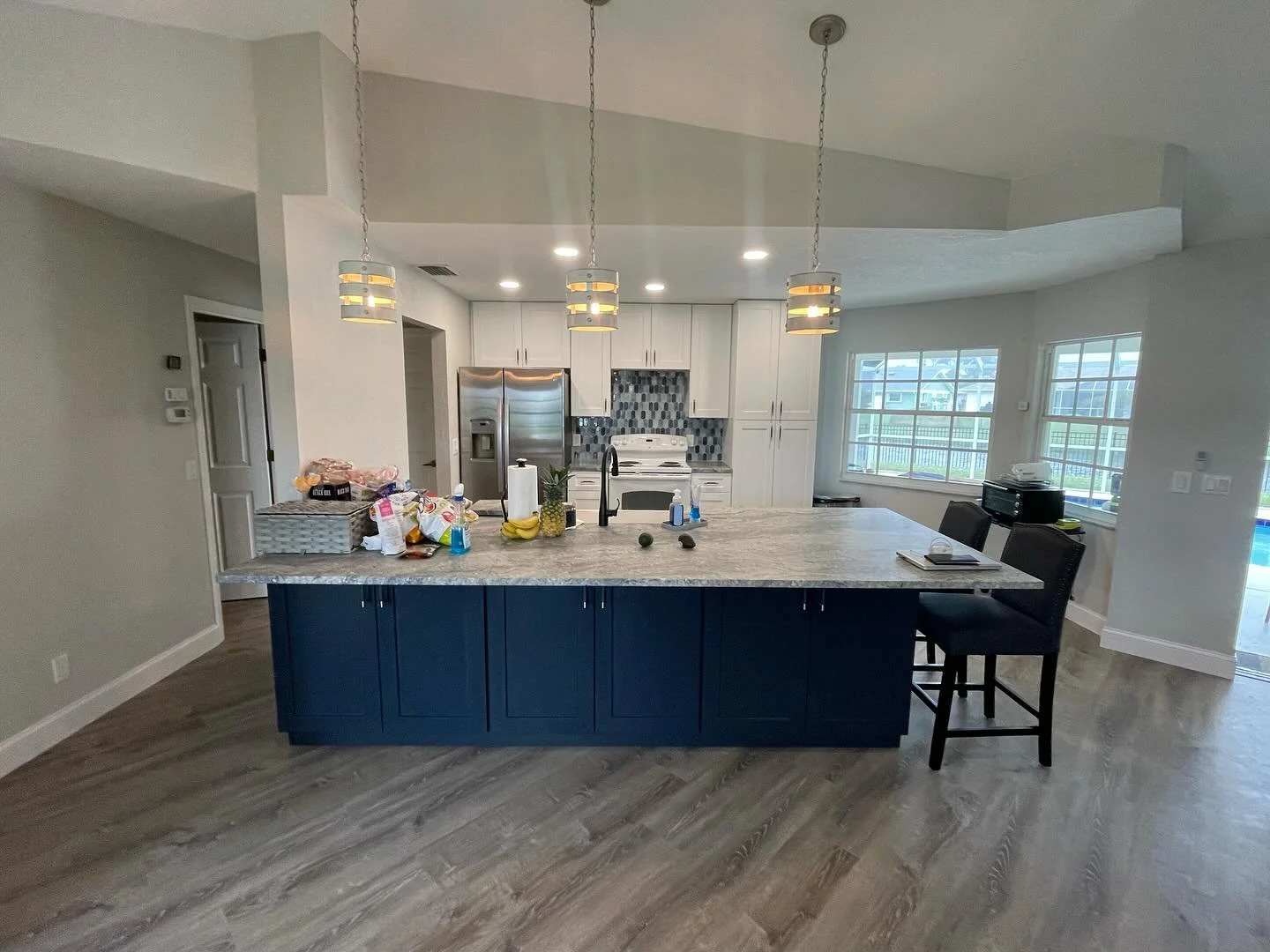 Modern kitchen with white cabinets, stainless steel refrigerator, white stove, and a large island with a marble countertop. The island has navy blue cabinets and is decorated with a pineapple, bananas, and various items. Three pendant lights hang abo