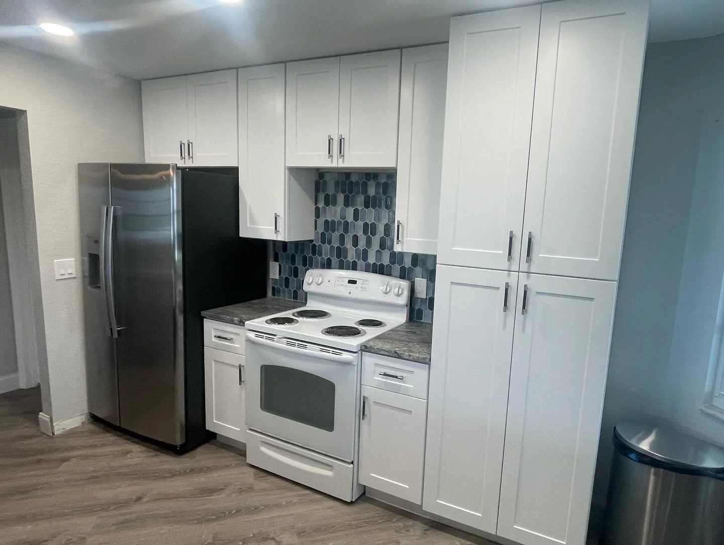 Modern kitchen with stainless steel refrigerator, white cabinets, granite countertops, stove, and a colorful hexagonal tile backsplash.