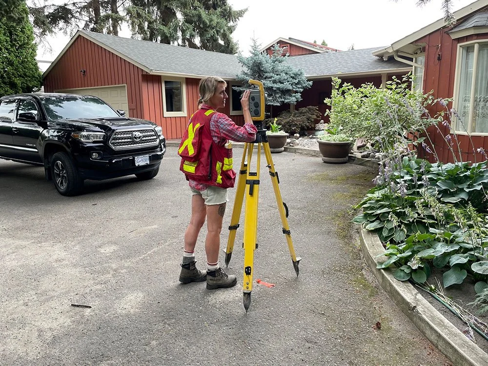 A woman in a red safety vest and shorts is operating a surveying instrument on a yellow tripod in a driveway. There is a black pickup truck and a red house with a garden to the right.