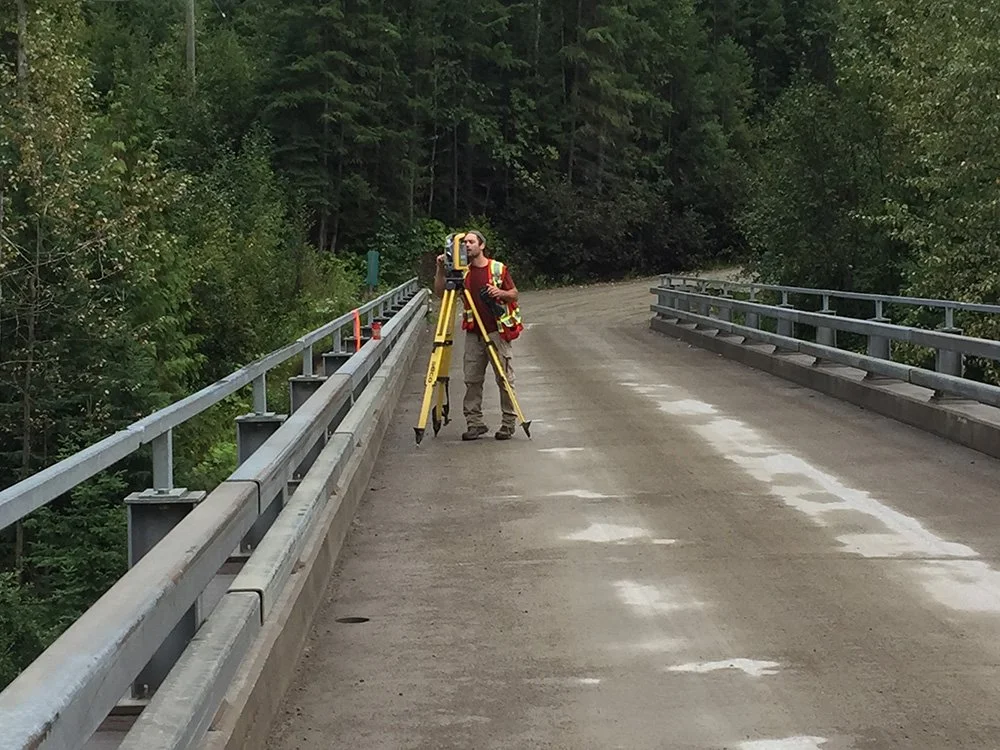 A surveyor standing on a bridge with trees in the background, using a total station and tripod to measure the area.