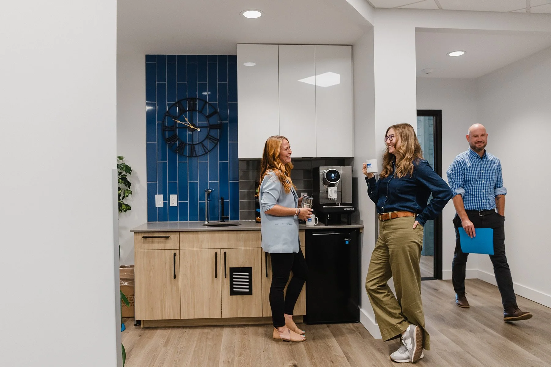 Three people standing and smiling in a modern office kitchen, with one woman holding a coffee mug, and two men holding notebooks, one with a tablet.
