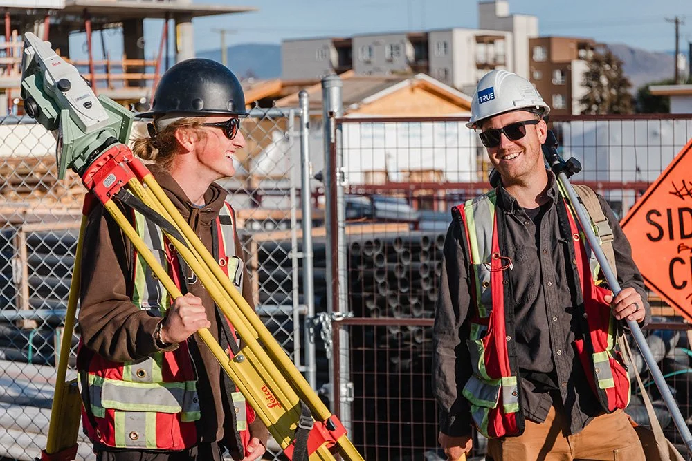 Two land surveyors, smiling and talking on a construction site. They are wearing safety helmets, sunglasses, and reflective safety vests, holding a yellow tripod, and holding a long pole.