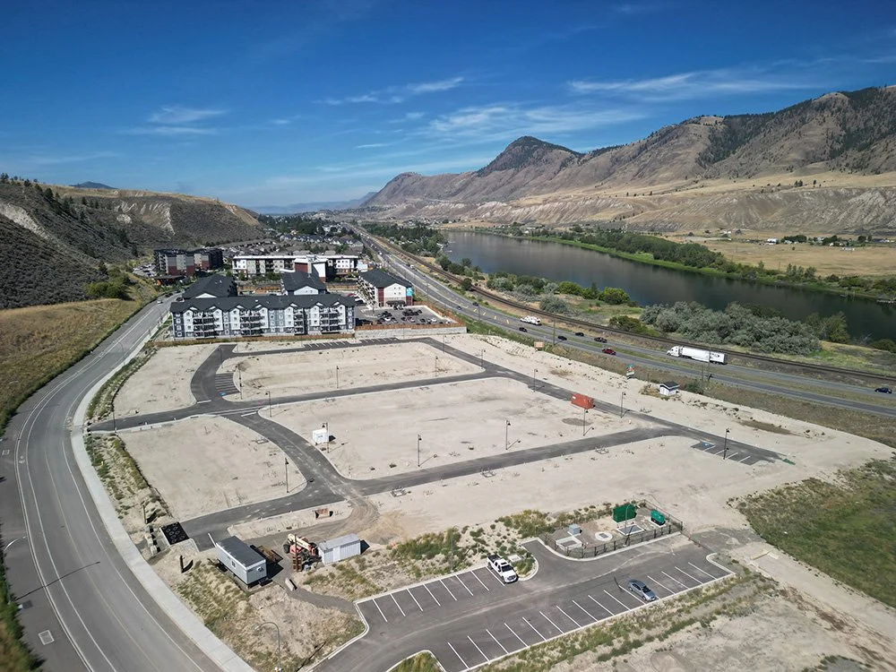 An aerial view of a construction site with cleared land, paved roads, and a few parked vehicles, situated near a river with a mountainous landscape in the background under a partly cloudy sky.