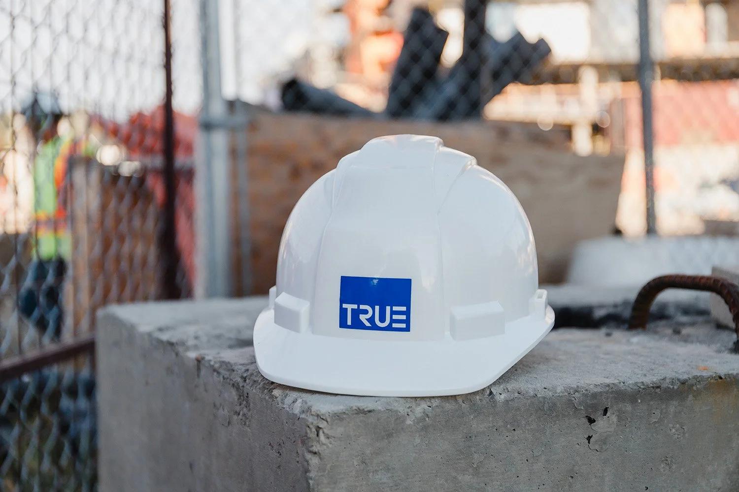 Construction safety helmet with a blue 'TRUE' logo resting on a concrete surface at a construction site.