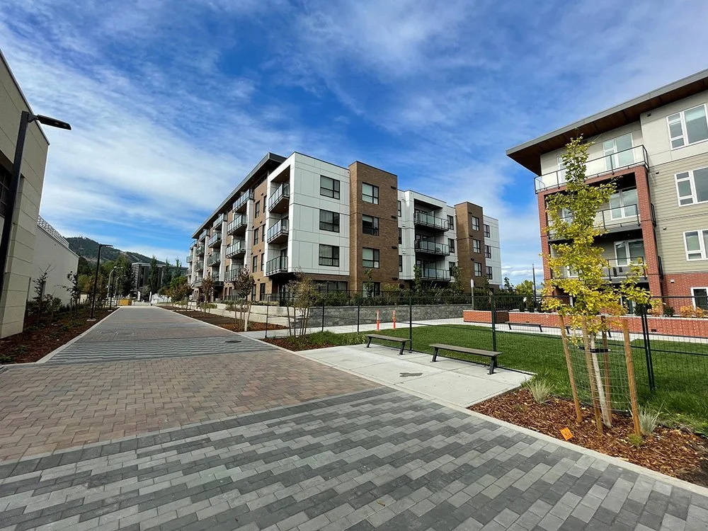 A modern residential apartment complex with multiple buildings, a paved walkway, benches, landscaped areas, and young trees under a partly cloudy sky.