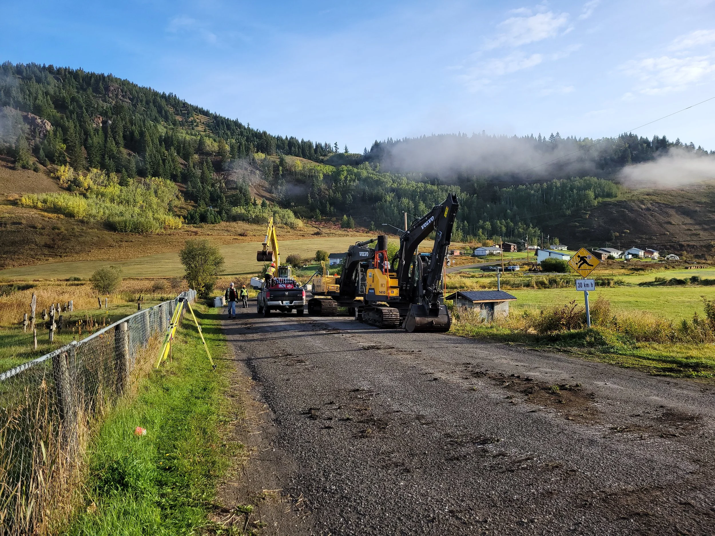 Construction site on a rural dirt road with excavators, a pickup truck, and workers, surrounded by green fields and hills with trees, under a blue sky with some clouds.