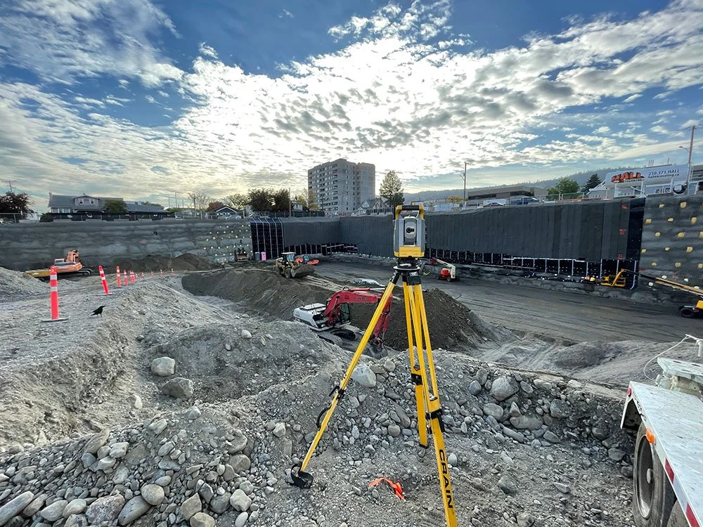 Construction site with heavy machinery, surveying equipment, and dirt mounds under a partly cloudy sky.