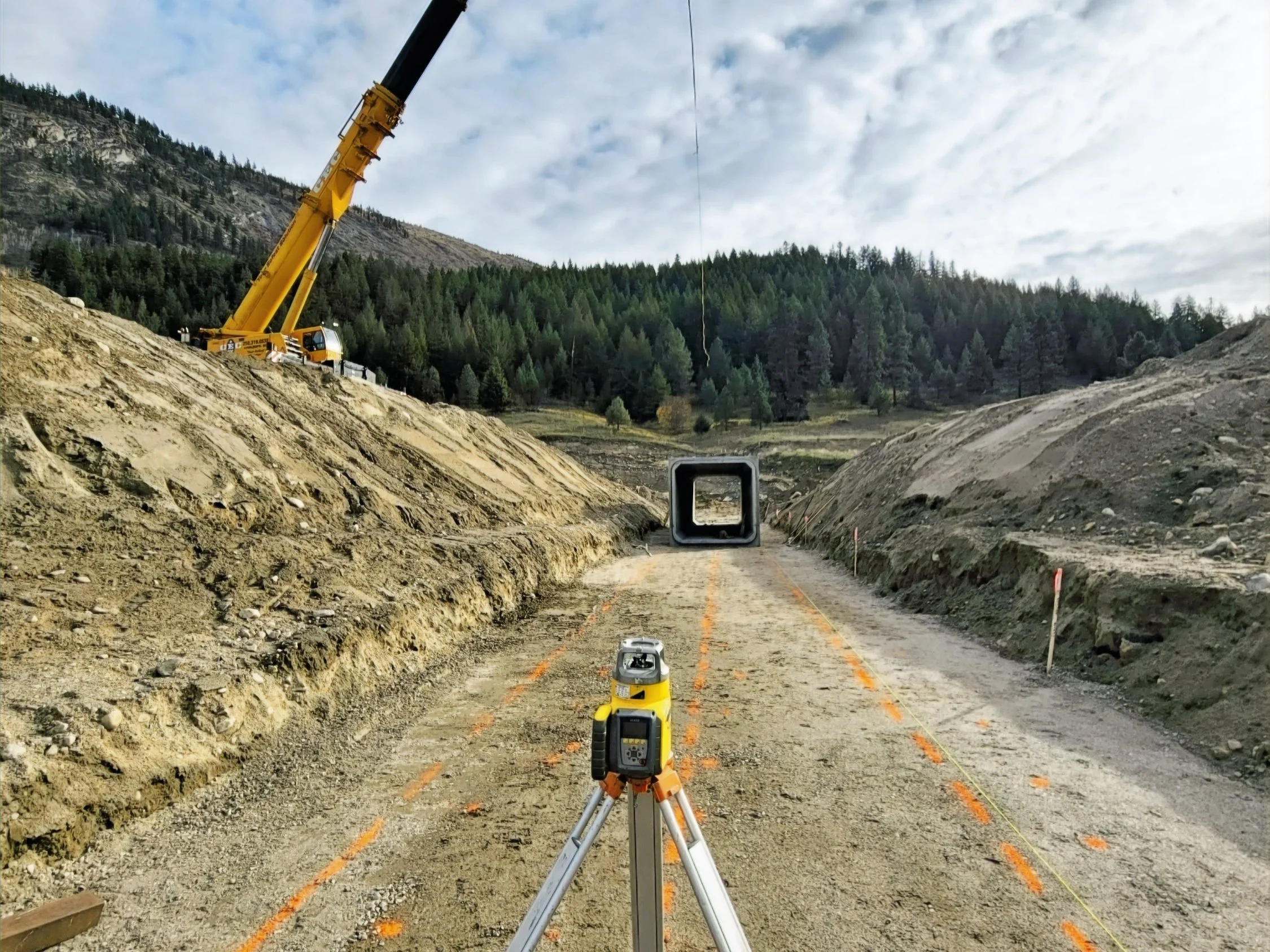 Construction site on a dirt road in a mountainous area with trees in the background. A yellow crane is on the left side, and surveying equipment is positioned in the foreground, facing a square tunnel in the distance.
