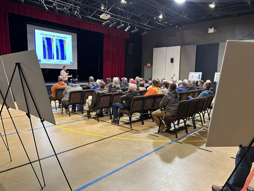 A speaker presenting on climate change hydrogen graphs to an audience seated in a theater with a stage and large screen.