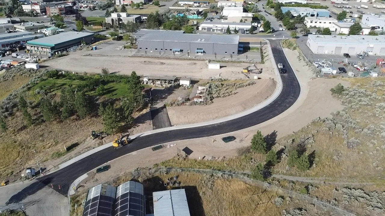 An aerial view of road construction in a suburban area, showing a newly paved winding road under development with construction equipment and barriers around it.