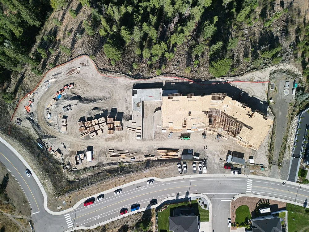 Aerial view of a construction site with a large building framework, construction materials, and equipment, surrounded by trees and roads.