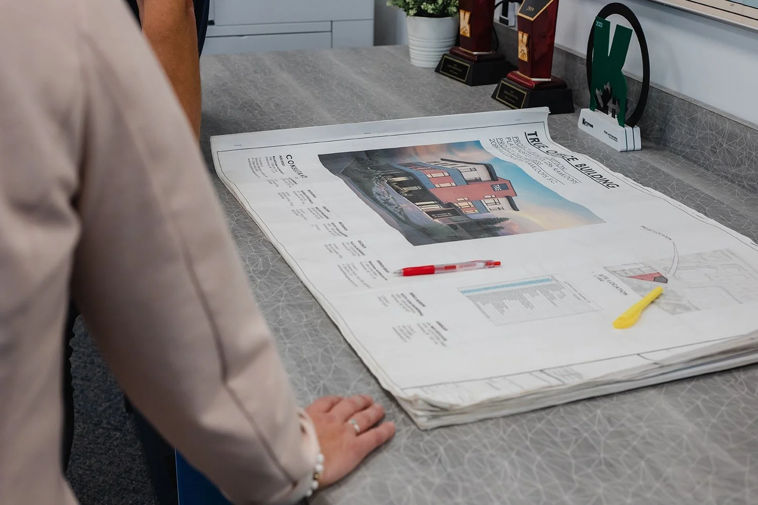 Architectural blueprint of a building on a large table with pens, trophies, and a potted plant in the background.