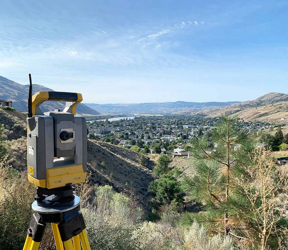 Surveying instrument on a tripod overlooks a wide valley with a river, houses, hills, and mountains under a clear blue sky.
