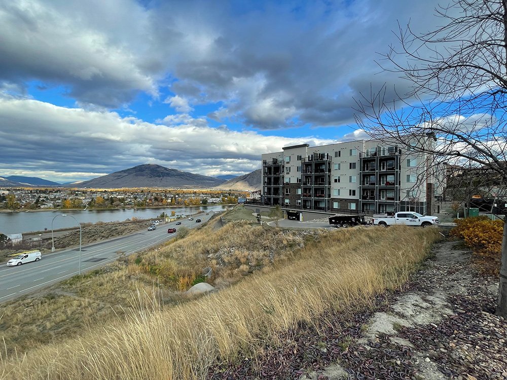 A cityscape with a river, a highway with cars, a modern apartment building, and mountains in the background under a partly cloudy sky.
