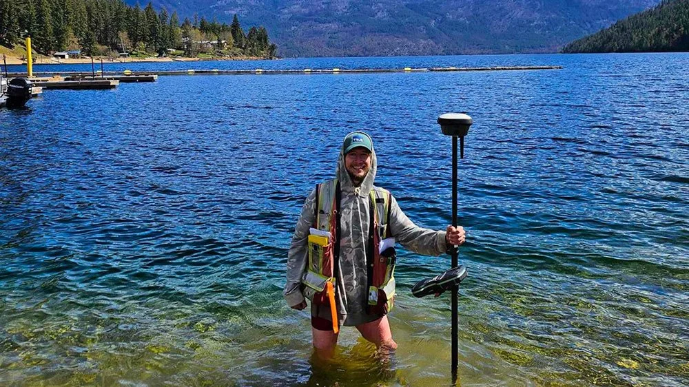 A person standing in clear lake water, holding a surveying tool, wearing a hoodie, vest, and shorts, with mountains and trees in the background.