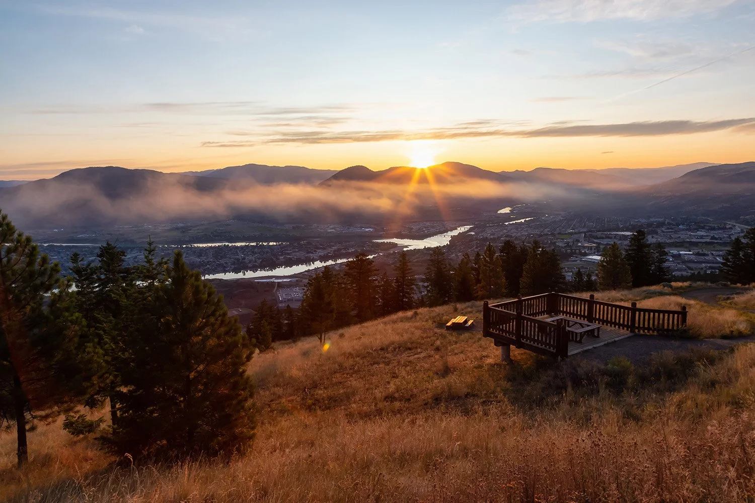 Sunset over a valley with mountains, river, trees, and a wooden viewing platform in Kamloops BC