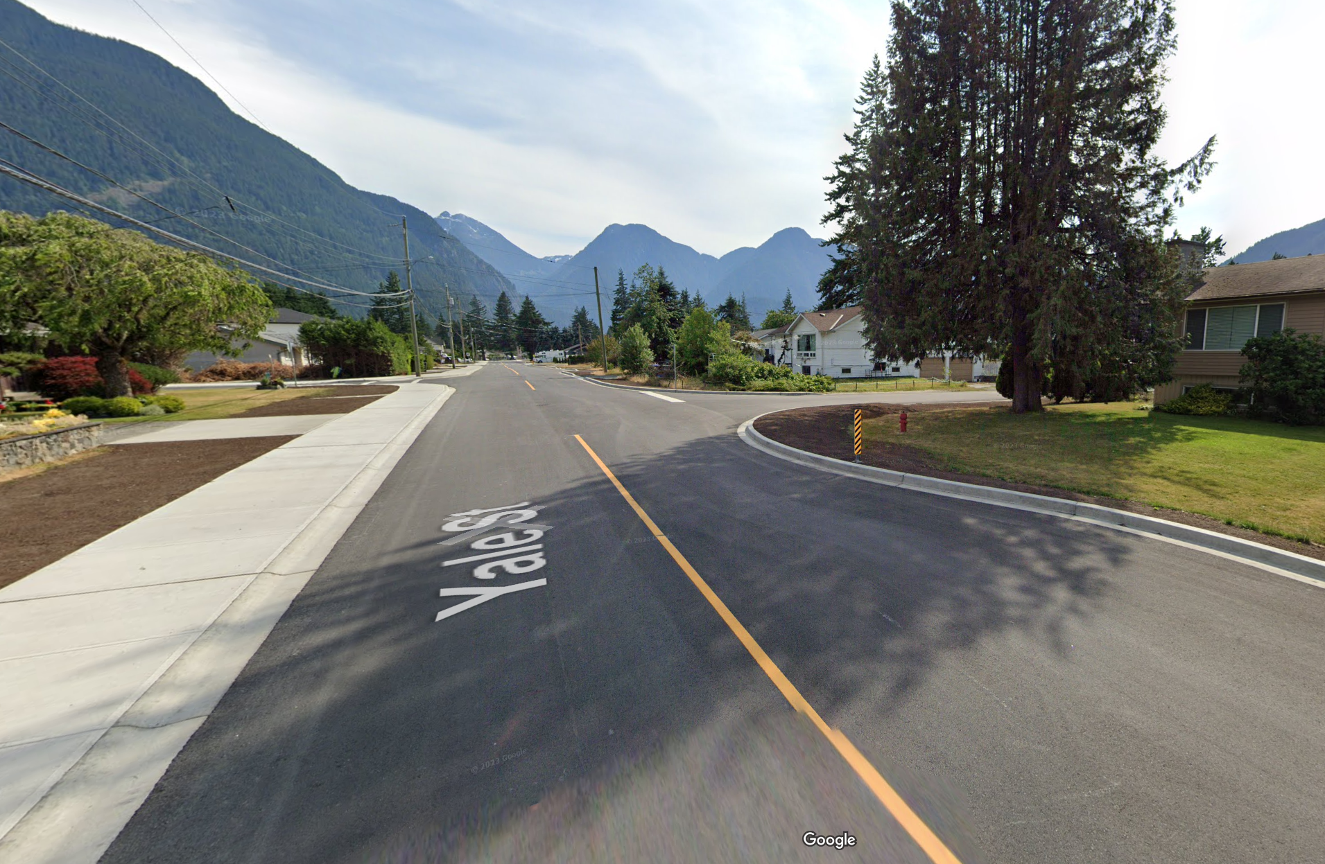 A peaceful residential street with houses, trees, and mountains in the background. The road is asphalt with a yellow dividing line and the sidewalk is concrete.