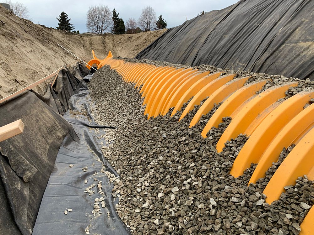 Construction site with a large trench reinforced with orange plastic or metal barriers and covered with black geotechnical fabric, filled with gravel or crushed stone.