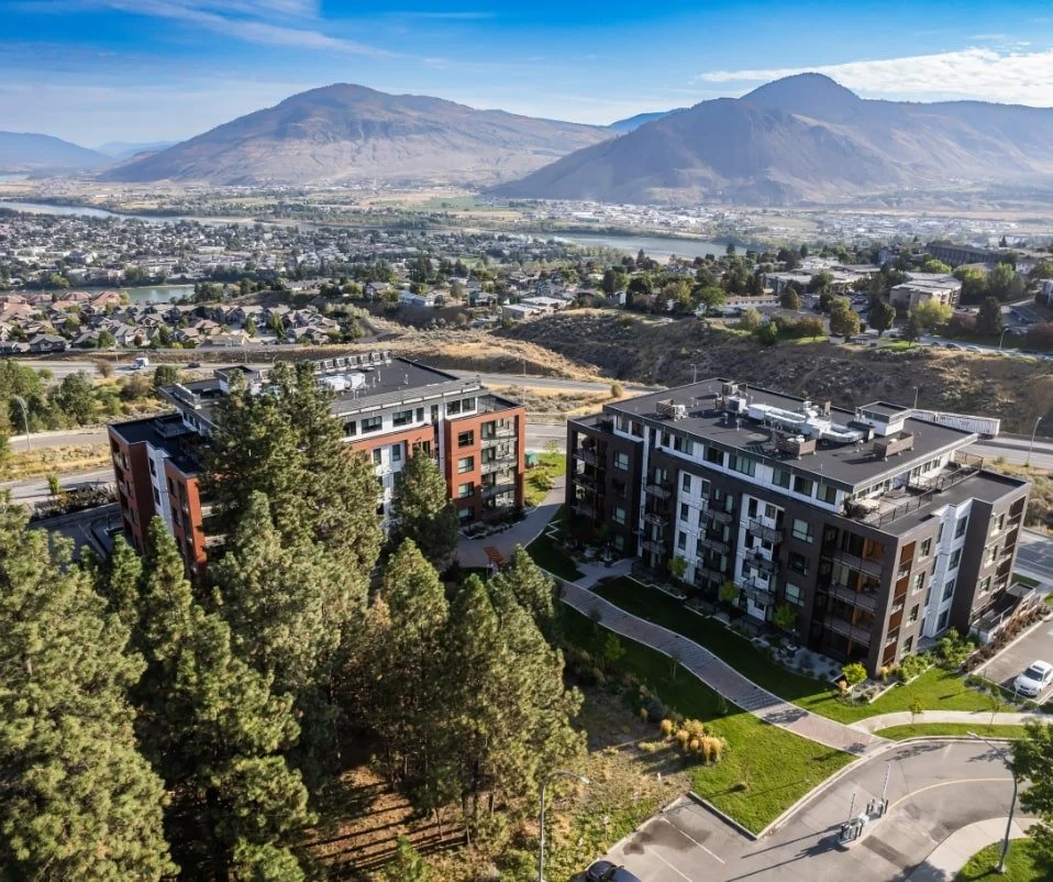 Aerial view of modern apartment buildings surrounded by trees with mountains in the background, residential area, and clear blue sky.