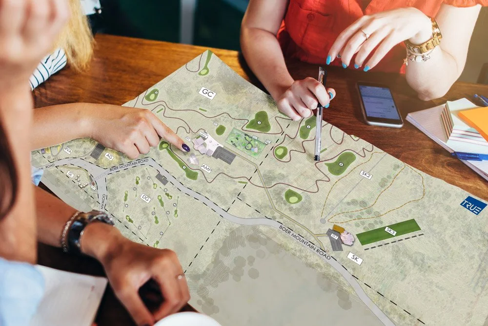 People reviewing a detailed map or site plan on a table, with one person pointing and another holding a pen, surrounded by notebooks and a smartphone.