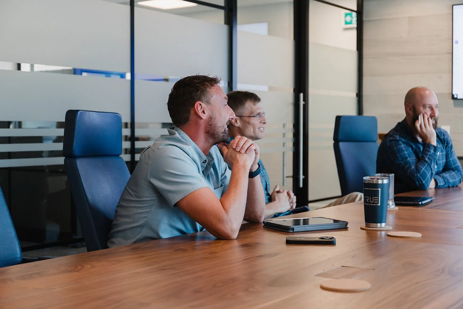 Three men sitting at a conference table in a modern office, engaging in a meeting or presentation, smiling and listening attentively.