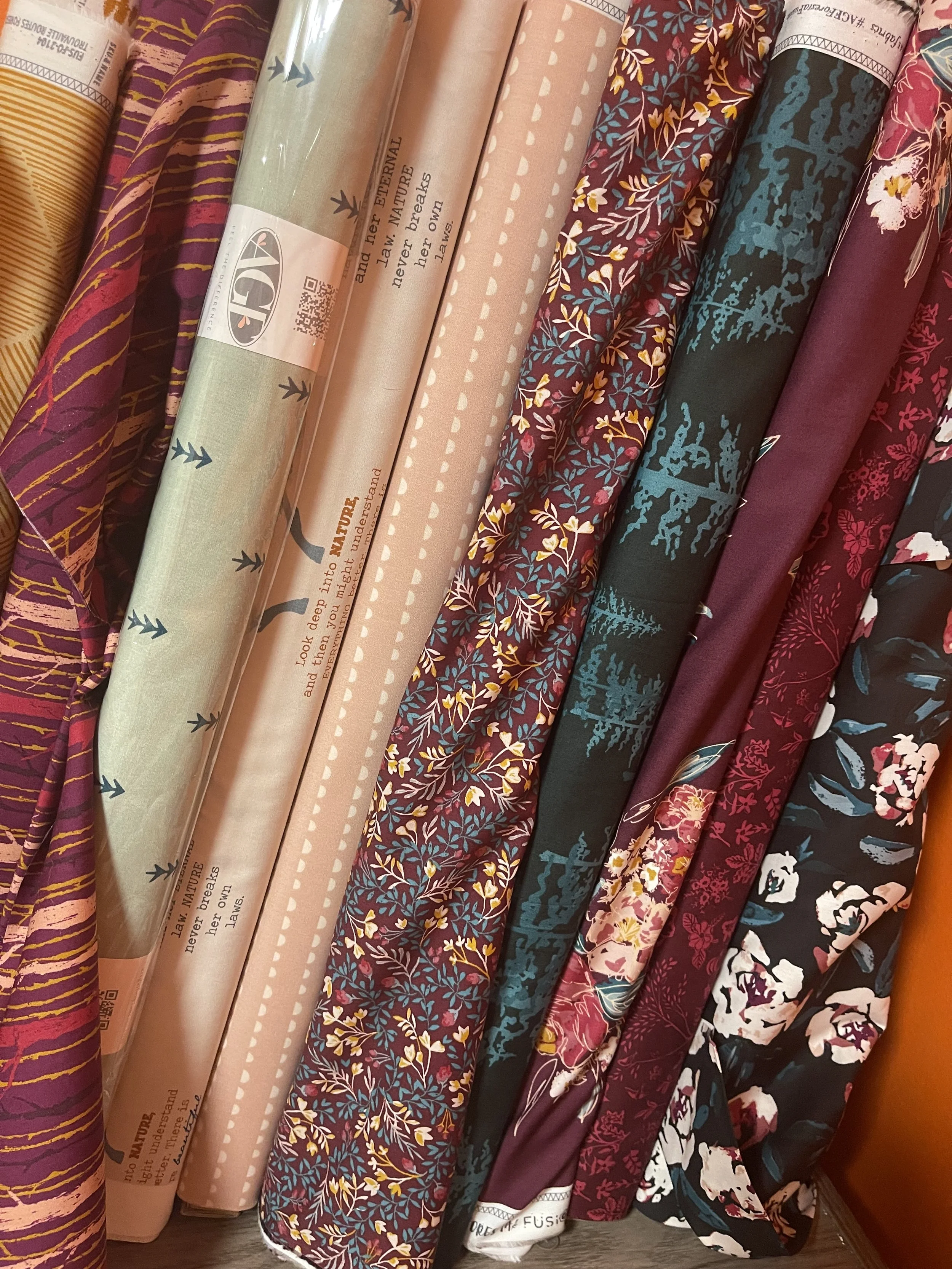 Stack of various patterned rolls of fabric in different colors including purple, pink, green, red, and black, with floral and nature-inspired designs.
