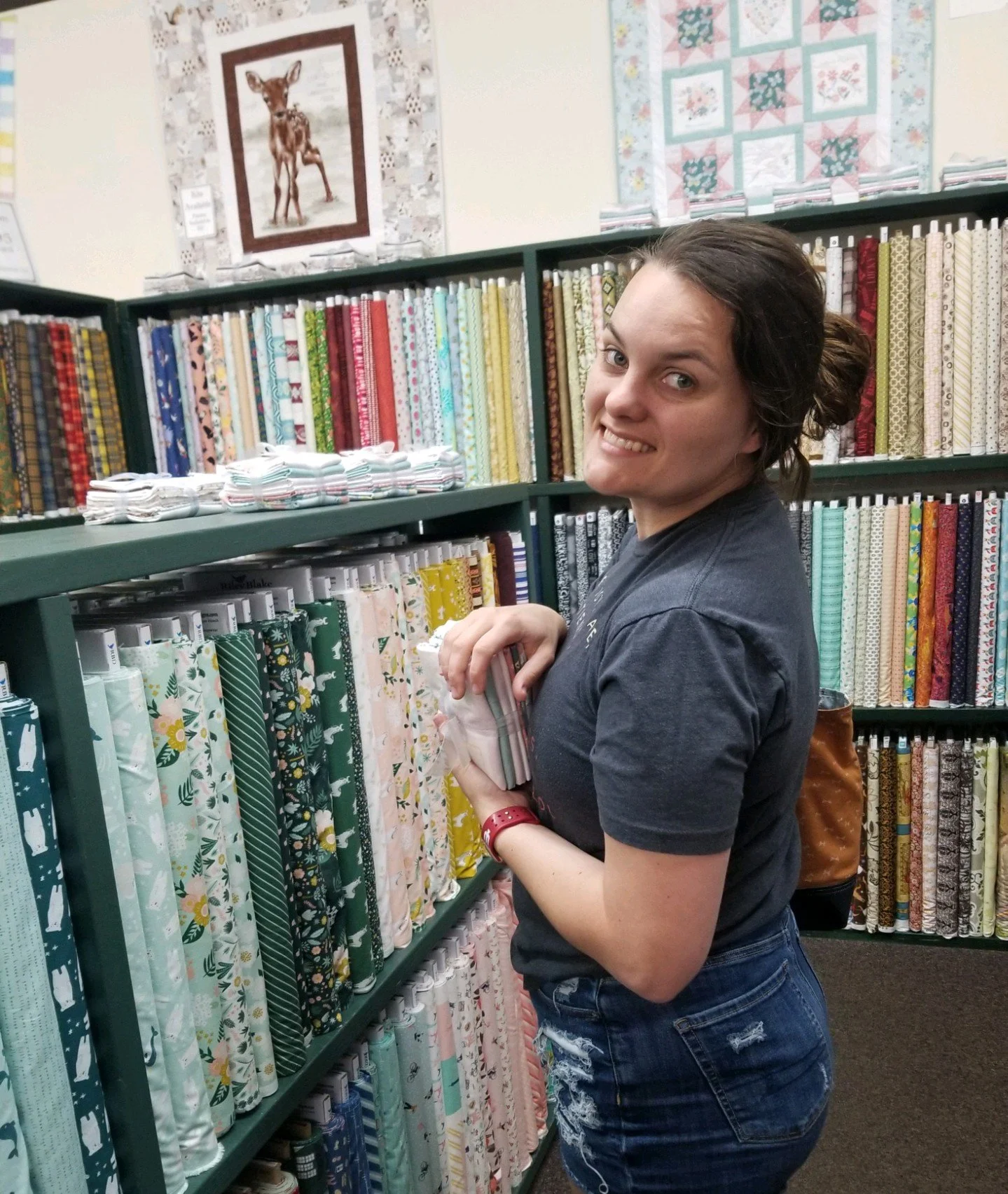Woman shopping for fabric in a store, holding multiple swatches and smiling at the camera with fabric bolts on shelves in the background.