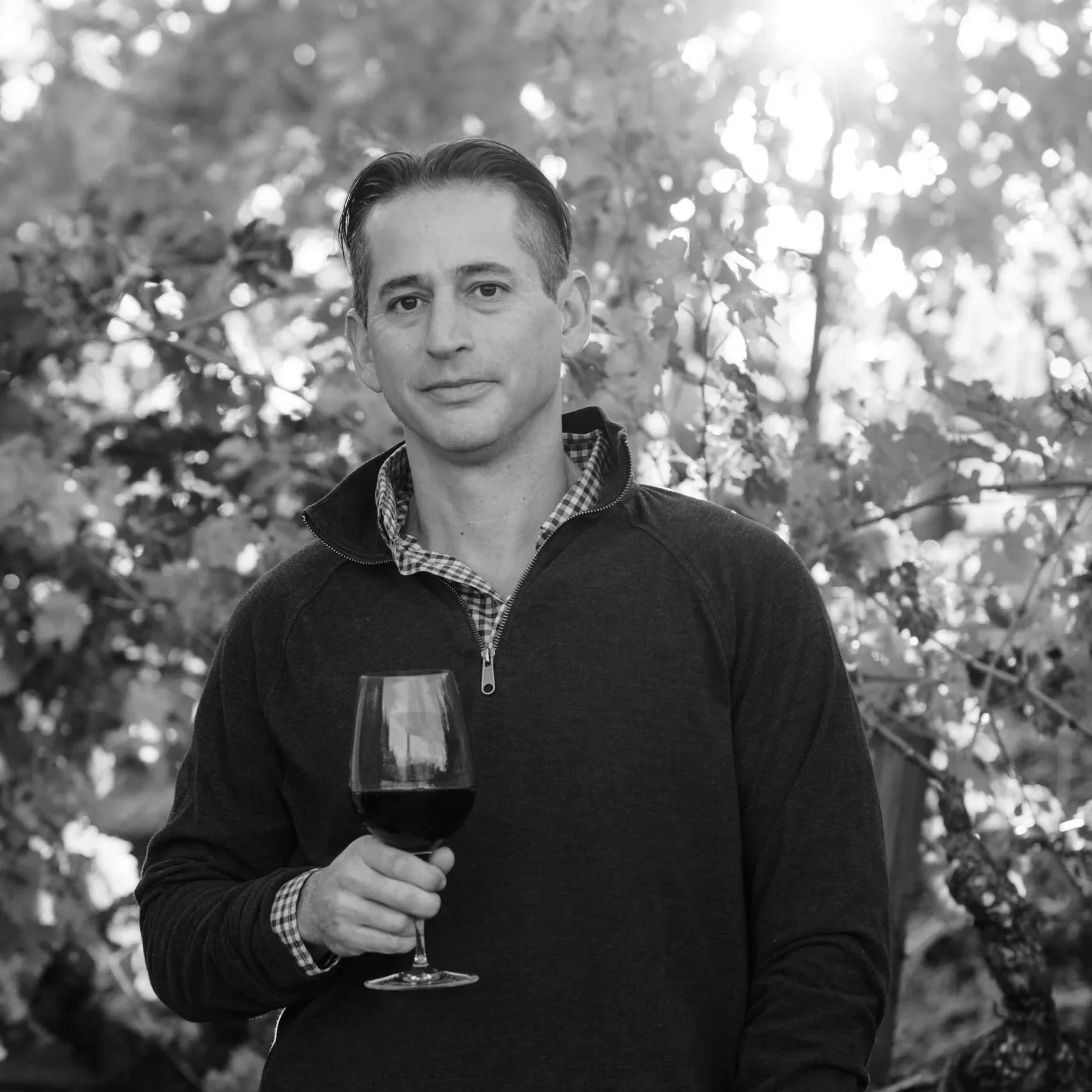 A man in a dark zip-up sweater holding a glass of red wine outdoors with trees and sunlight in the background.