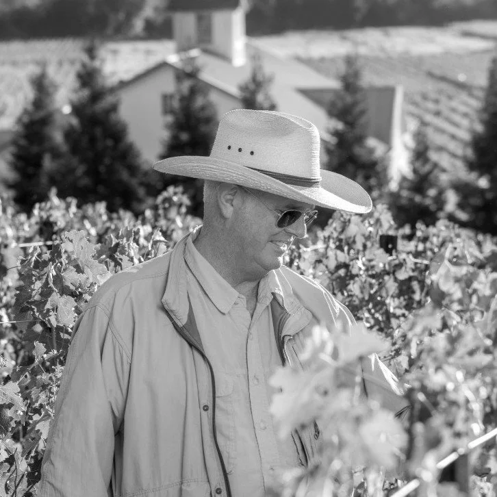 Man in a wide-brimmed hat and sunglasses stands in a vineyard with grapevines in the background, looking down.