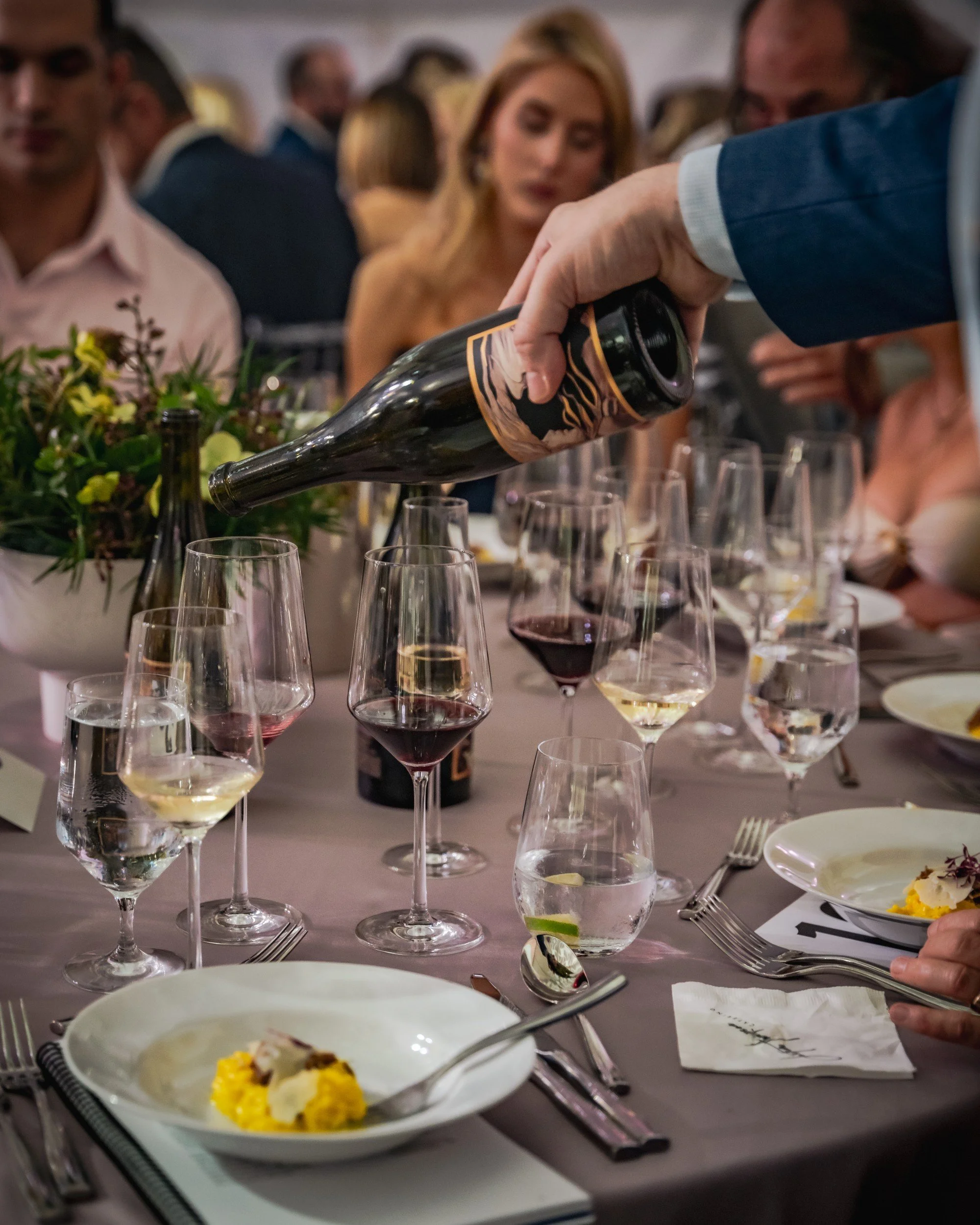 Person pouring red wine into a glass at an elegant dinner party with multiple wine glasses, plates, silverware, and a floral centerpiece on the table.