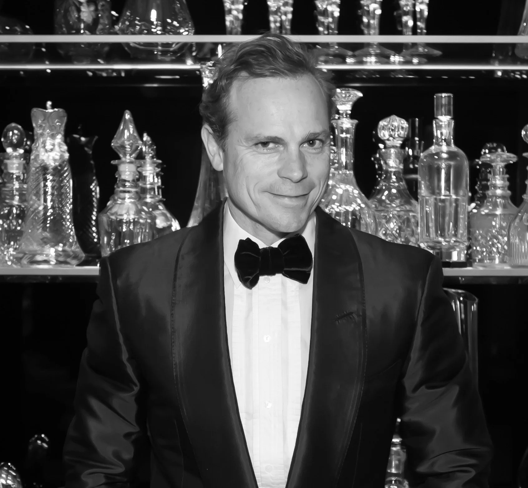 A man in a tuxedo with a bow tie standing in front of a shelf filled with glass decanters.