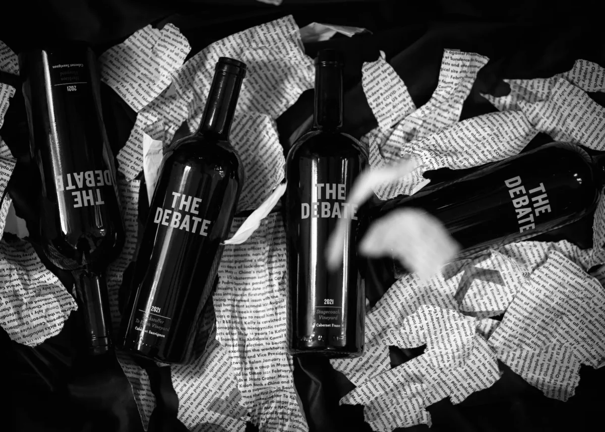 Four black bottles labeled "The Debate" surrounded by crumpled newspaper and fabric.