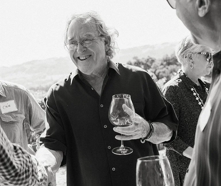 Smiling man with glasses holding a wine glass at an outdoor social gathering.