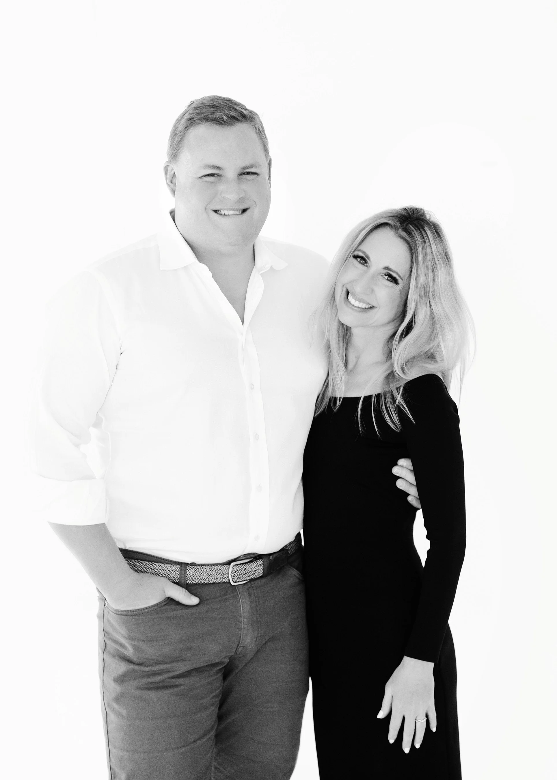 A black and white photo of a smiling man and woman standing close together, with the woman's hand on the man's waist, against a plain white background.