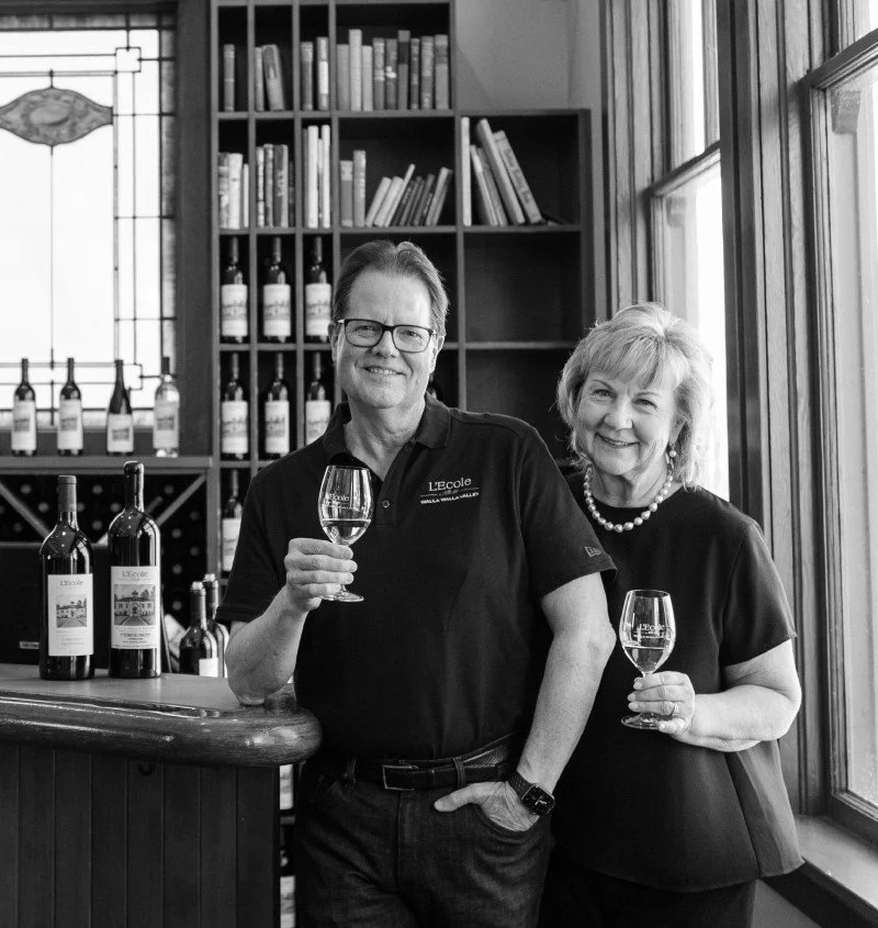 A man and woman smiling and holding wine glasses inside a wine bar or tasting room, with wine bottles and a bookshelf in the background.