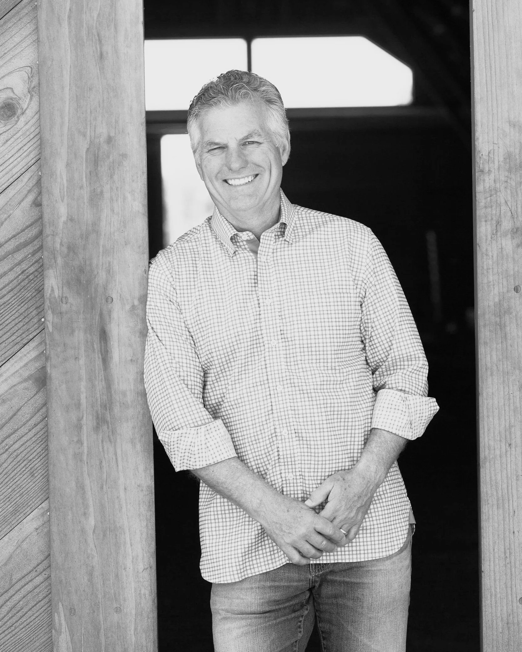 A black and white photo of a smiling man with short, light-colored hair, wearing a checkered button-up shirt with sleeves rolled up, standing in a wooden doorway.