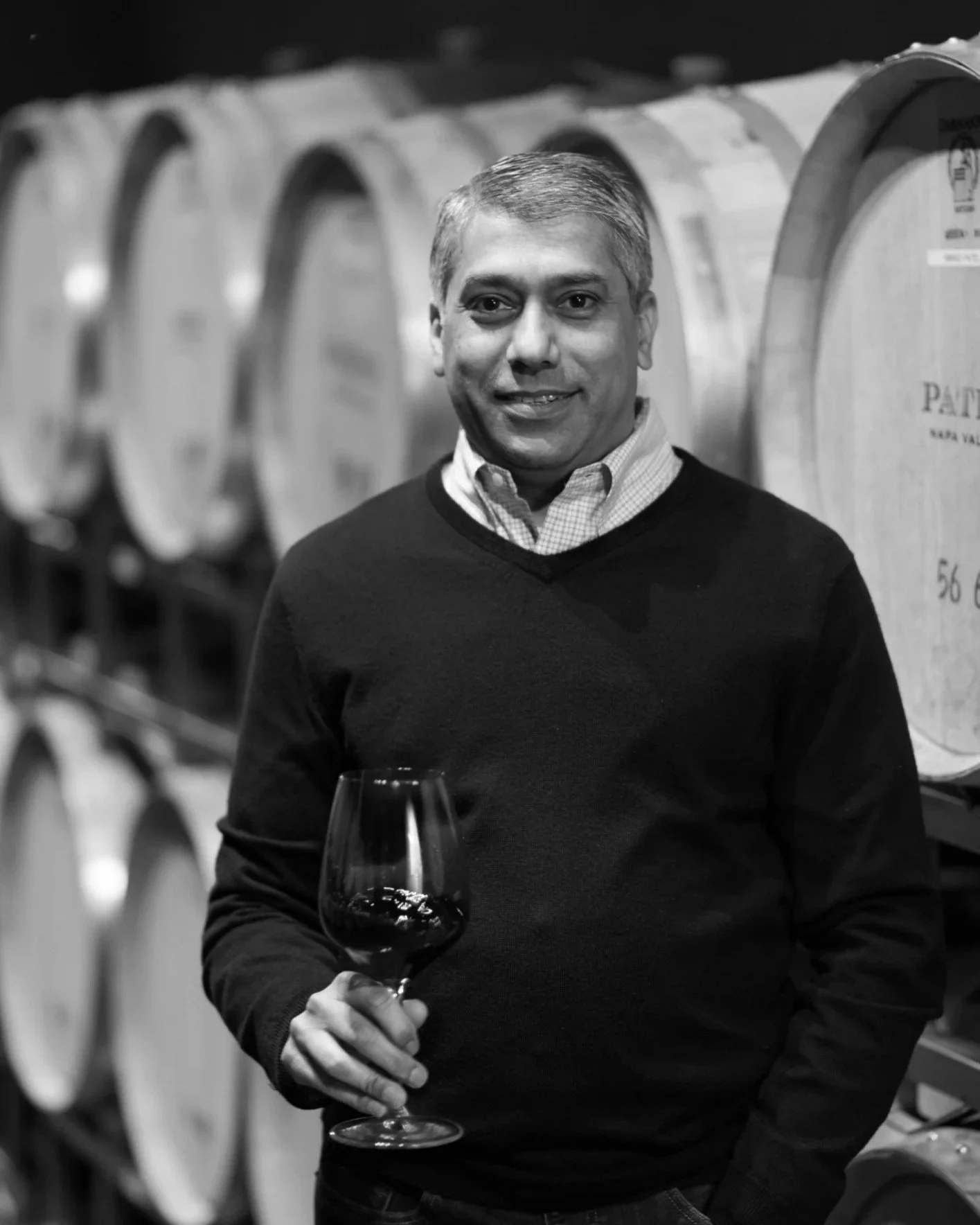 A man holding a glass of red wine in a wine cellar with rows of wine barrels in the background.