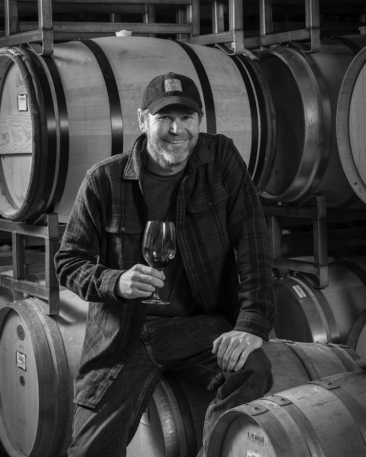 A smiling man with a beard wearing a baseball cap and plaid shirt, holding a glass of wine, sitting on a wooden barrel in a winery surrounded by wine barrels.