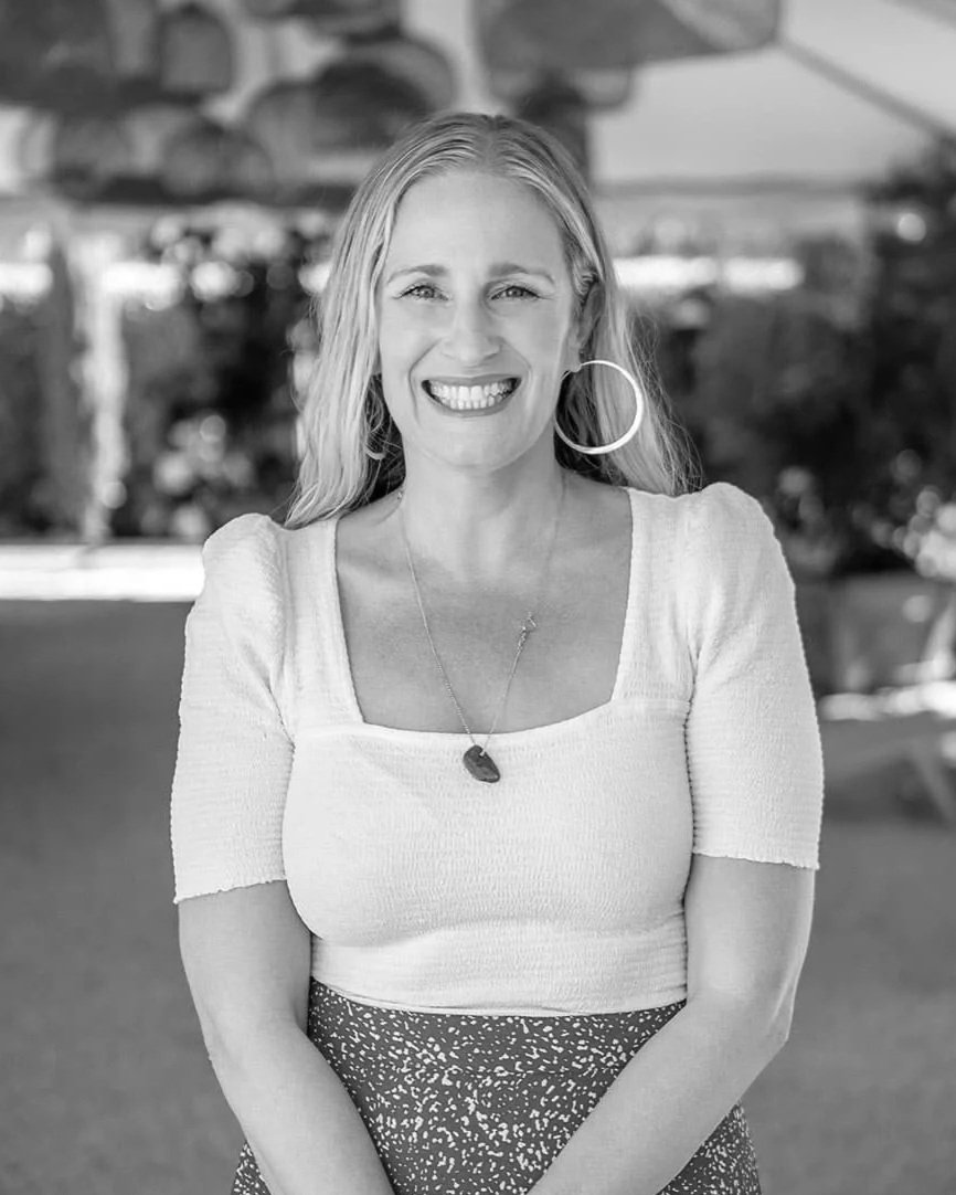 Black and white photo of a smiling woman with long hair, wearing a short-sleeved top, large hoop earrings, a necklace with a pendant, and a patterned skirt, standing outdoors.