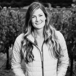 A woman with long, wavy hair smiling outdoors, wearing a light-colored jacket.