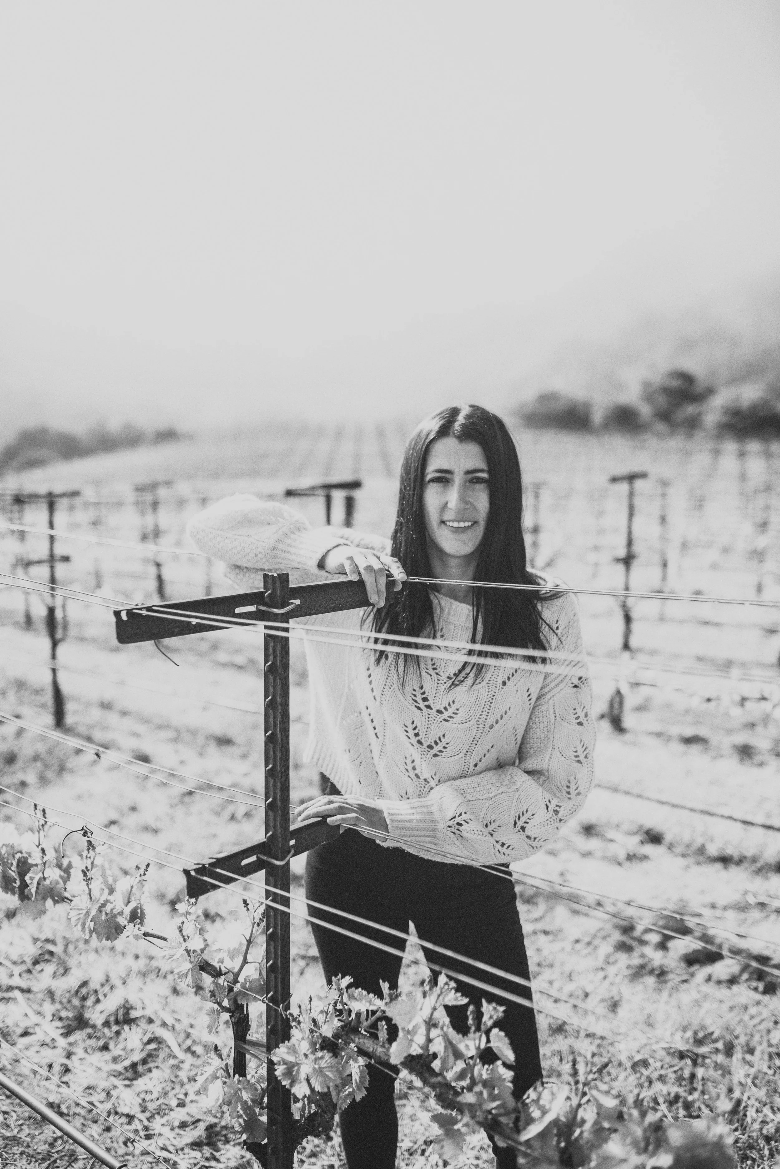 A woman with long dark hair smiling while standing in a vineyard, holding onto a wire trellis.