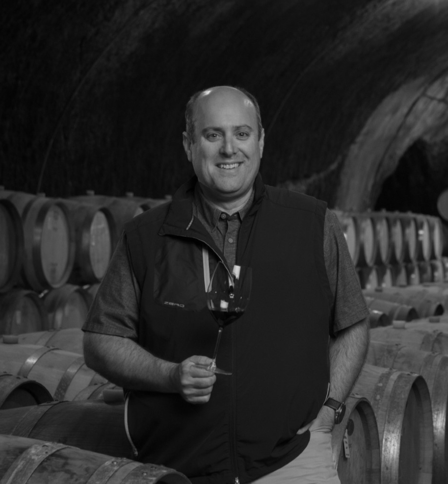 A smiling man holding a glass of red wine in a wine cellar surrounded by wooden barrels.
