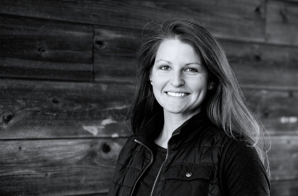 Black and white portrait of a woman with long hair, smiling, wearing a dark jacket, standing against a wooden background.