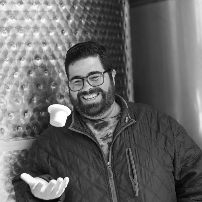 A man with glasses, a beard, and a smile, wearing a jacket, tossing a small white cup into the air.